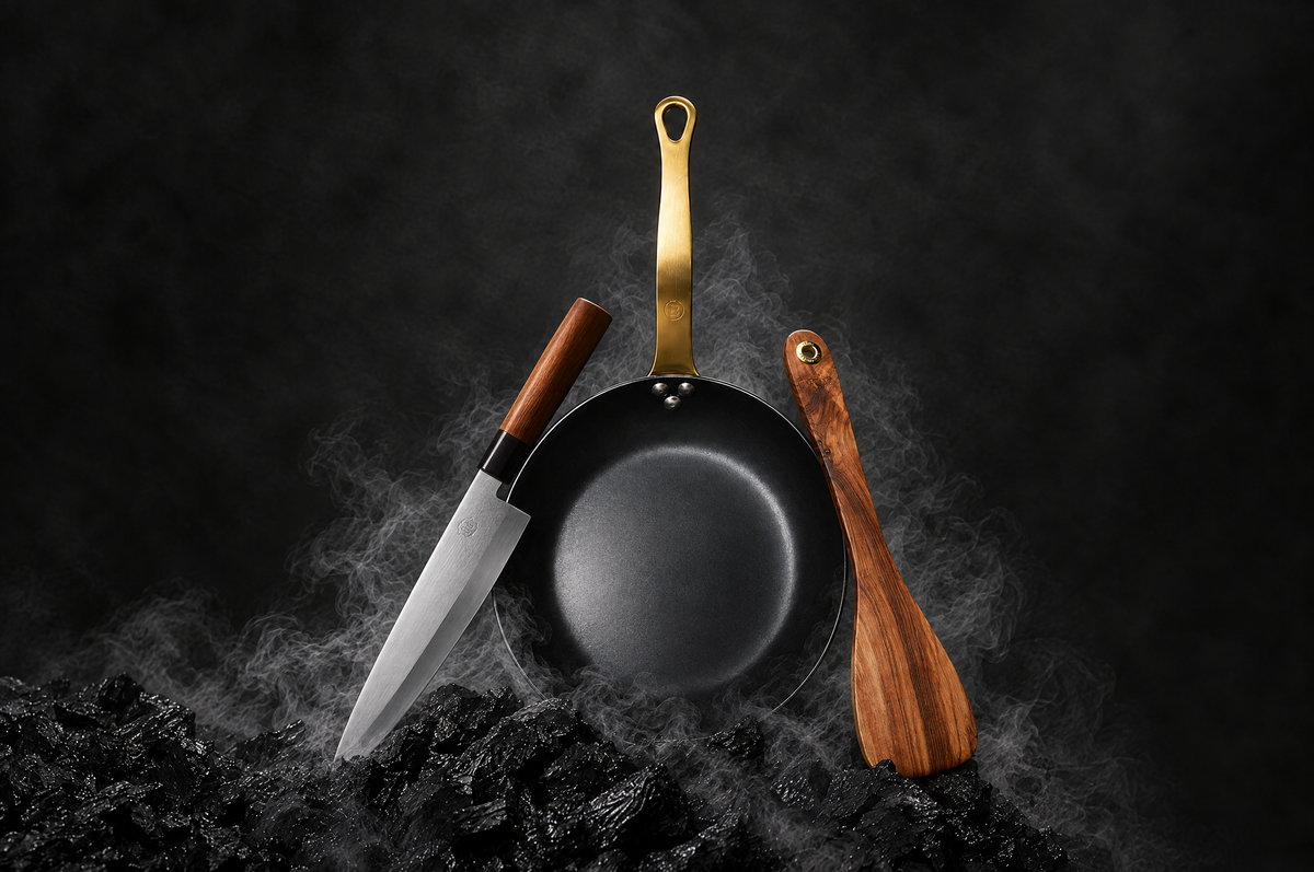 Padella NeroSteel, coltello giapponese e spatola in ulivo tra fumo e carbone — immagine generata con AI